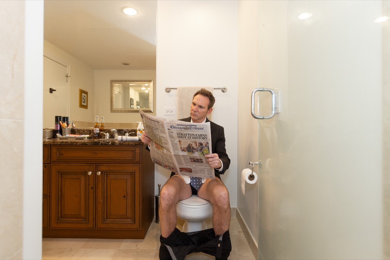 Jon reads a newspaper while seated on a toilet in a bathroom.